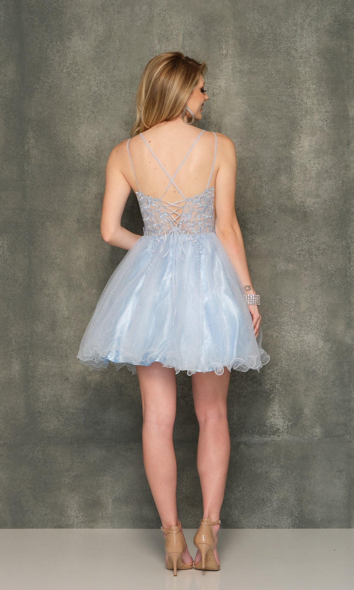 A woman faces away, wearing the Sheer-Bodice Ice Blue Short Homecoming Dress 10844 featuring a lace-up back, spaghetti straps, and fit-and-flare design with nude heels against a plain gray background.